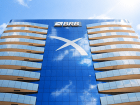 Fachada do edifício do BRB em Brasília, vista em ângulo baixo e mais iluminada, com vidro azul refletindo o céu, simbolizando modernização e serviços financeiros.