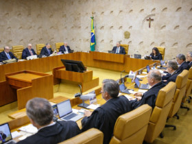 Ministros do Supremo Tribunal Federal reunidos em sessão plenária, sentados em bancadas de madeira no plenário oficial, com bandeira do Brasil ao fundo e ambiente institucional formal.