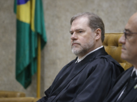 Magistrado do Supremo Tribunal Federal com toga preta, sentado em cadeira de couro no plenário, com bandeira do Brasil ao fundo e expressão séria durante julgamento.