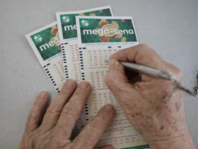Pessoa preenchendo bilhete da Mega-Sena com caneta em mesa clara, com vários volantes ao fundo prontos para conferência do sorteio.