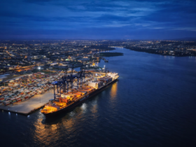 Vista aérea noturna de porto brasileiro com navio cargueiro, guindastes e contêineres iluminados, refletindo luzes na água e cidade ao fundo.