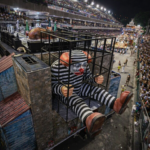 Alegoria de palhaço atrás das grades em carro alegórico durante desfile na Marquês de Sapucaí, com público nas arquibancadas à noite