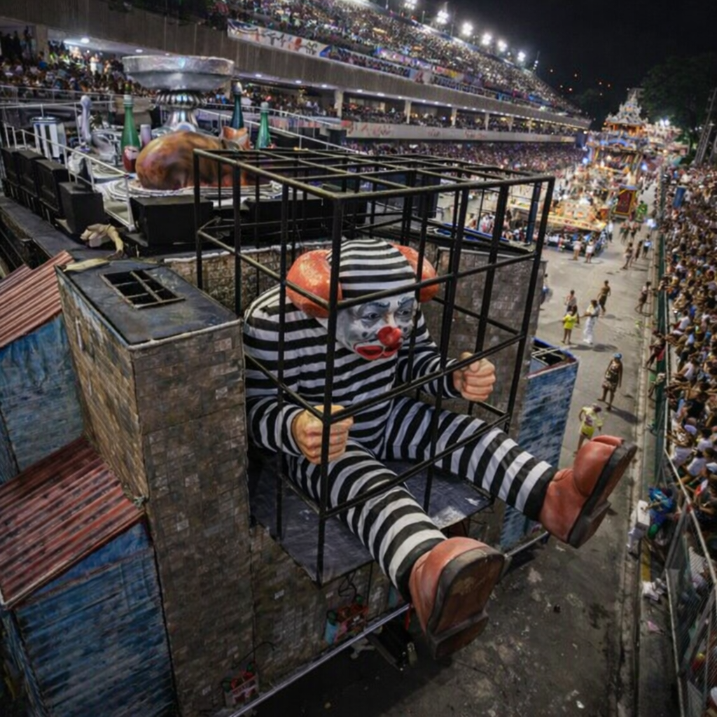 Alegoria de palhaço atrás das grades em carro alegórico durante desfile na Marquês de Sapucaí, com público nas arquibancadas à noite