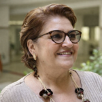 Ministra Cida Gonçalves sorrindo em retrato de perfil leve, usando óculos e colar artesanal, em ambiente institucional iluminado por luz natural