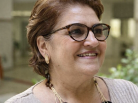 Ministra Cida Gonçalves sorrindo em retrato de perfil leve, usando óculos e colar artesanal, em ambiente institucional iluminado por luz natural