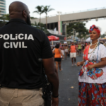 Agente da Polícia Civil patrulhando circuito do Carnaval de Salvador, com foliões ao redor e estrutura de camarotes ao fundo