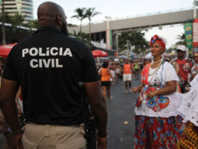 Agente da Polícia Civil patrulhando circuito do Carnaval de Salvador, com foliões ao redor e estrutura de camarotes ao fundo