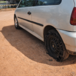 Veículo branco parado em estrada de terra com pneu traseiro completamente estourado e roda exposta sob luz do dia