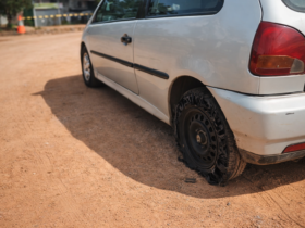 Veículo branco parado em estrada de terra com pneu traseiro completamente estourado e roda exposta sob luz do dia