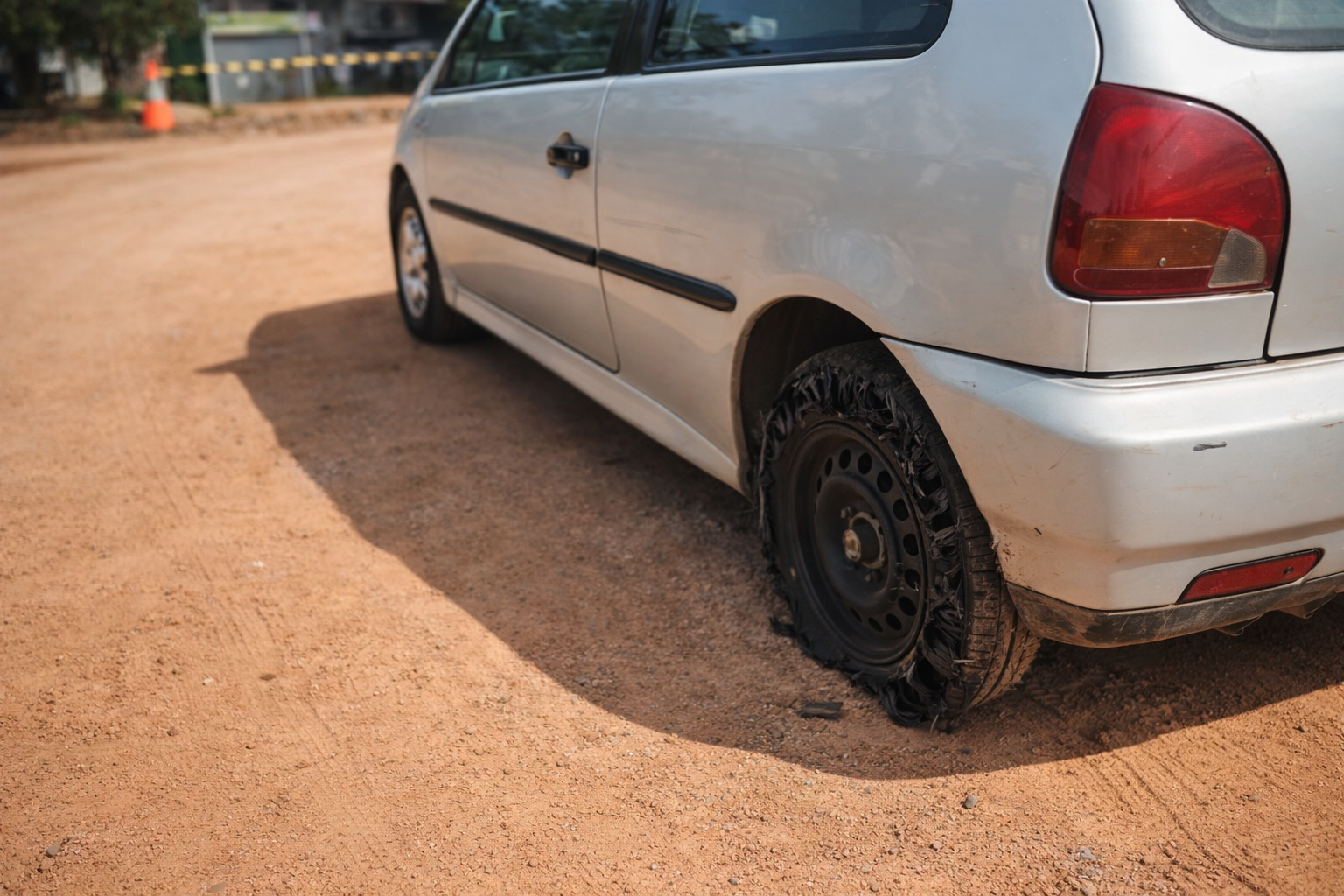 Veículo branco parado em estrada de terra com pneu traseiro completamente estourado e roda exposta sob luz do dia