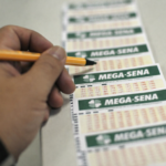 Pessoa segurando lápis enquanto marca volante da Mega-Sena sobre balcão, com sequência de bilhetes alinhados ao fundo