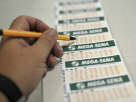 Pessoa segurando lápis enquanto marca volante da Mega-Sena sobre balcão, com sequência de bilhetes alinhados ao fundo