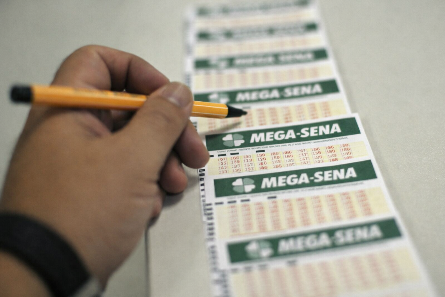 Pessoa segurando lápis enquanto marca volante da Mega-Sena sobre balcão, com sequência de bilhetes alinhados ao fundo