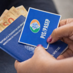Mãos segurando cartão do PIS/Pasep junto a carteira de trabalho e dinheiro em real, representando pagamento do abono salarial no Brasil
