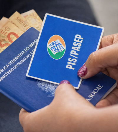 Mãos segurando cartão do PIS/Pasep junto a carteira de trabalho e dinheiro em real, representando pagamento do abono salarial no Brasil