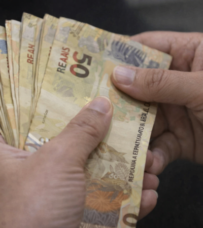 Mãos segurando e contando notas de 50 reais, representando pagamento, renda e movimentação financeira no Brasil