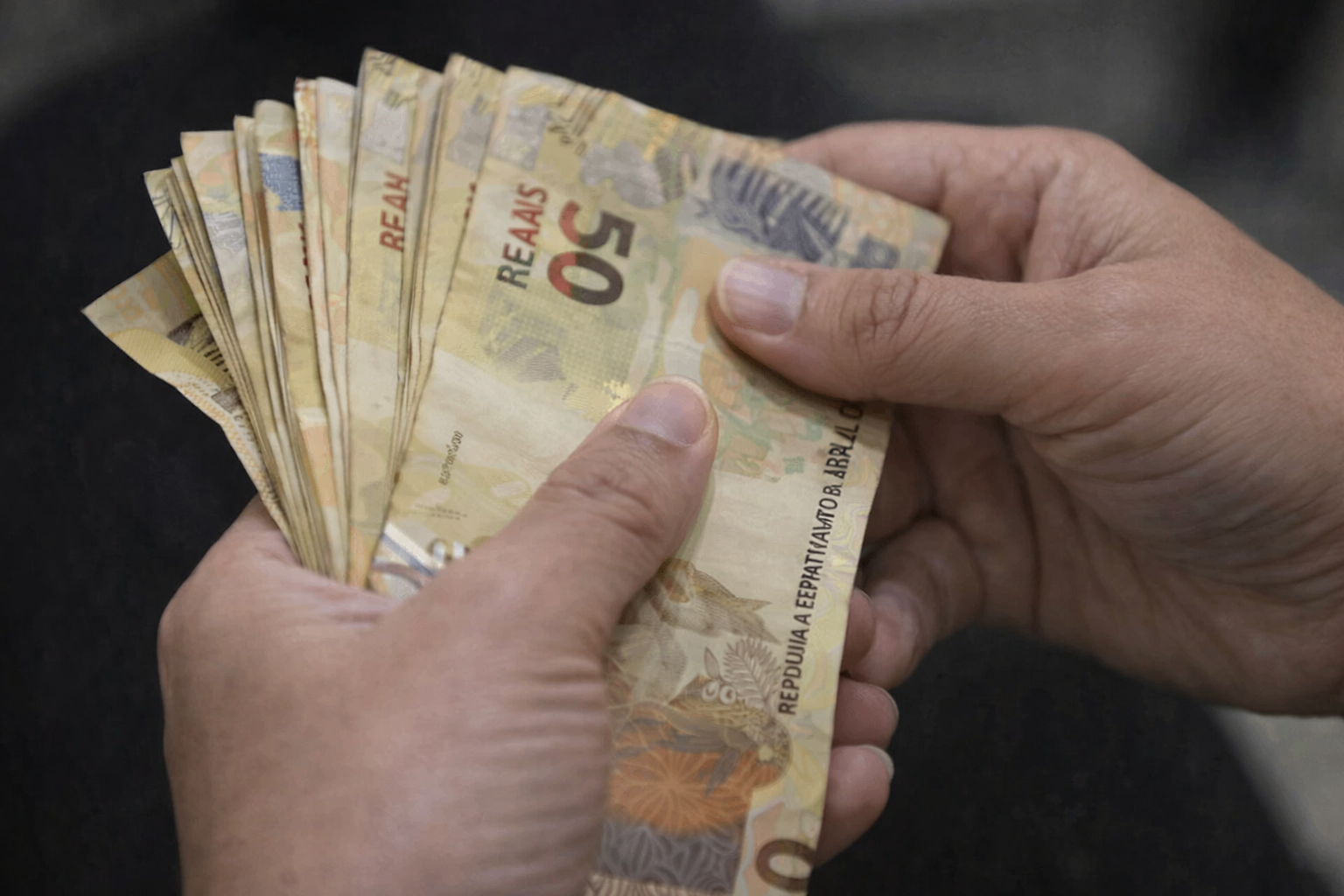 Mãos segurando e contando notas de 50 reais, representando pagamento, renda e movimentação financeira no Brasil