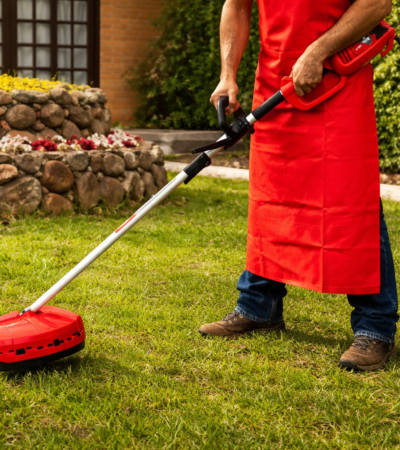 Pessoa operando aparador elétrico de grama em jardim residencial, realizando manutenção do gramado no verão.
