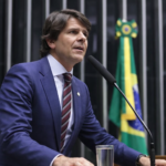 Deputado em perfil discursando na tribuna da Câmara dos Deputados com bandeira do Brasil ao fundo em Brasília
