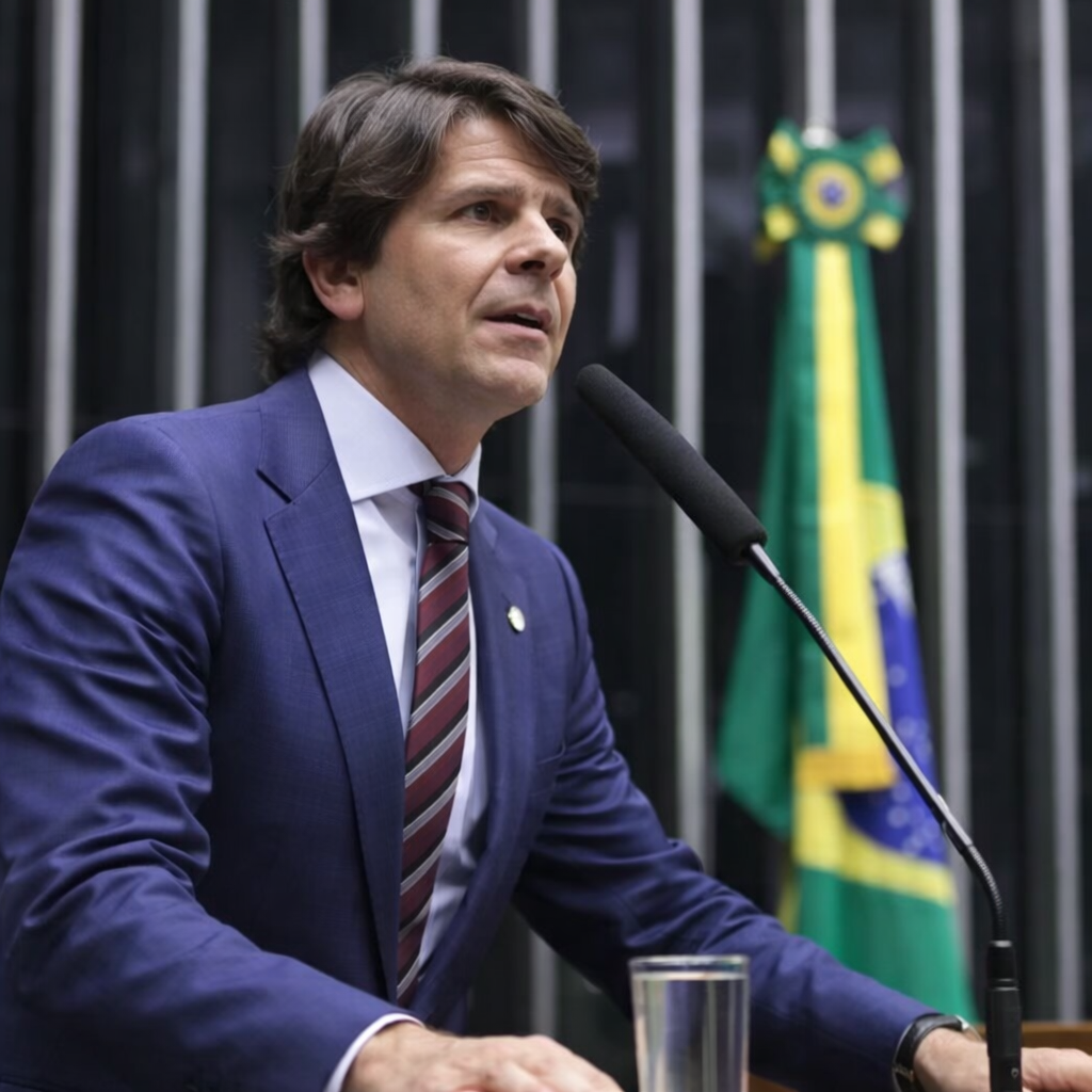 Deputado em perfil discursando na tribuna da Câmara dos Deputados com bandeira do Brasil ao fundo em Brasília