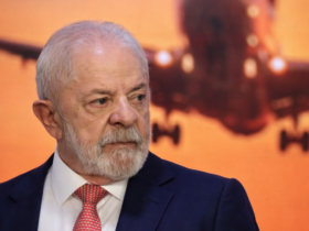 Luiz Inácio Lula da Silva em plano médio durante evento oficial, vestindo terno azul e gravata vermelha, com imagem de aeronave iluminada ao fundo.