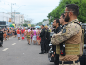 Agentes de segurança monitoram bloco de rua durante o carnaval, com viaturas ao fundo e foliões participando do evento em área urbana.