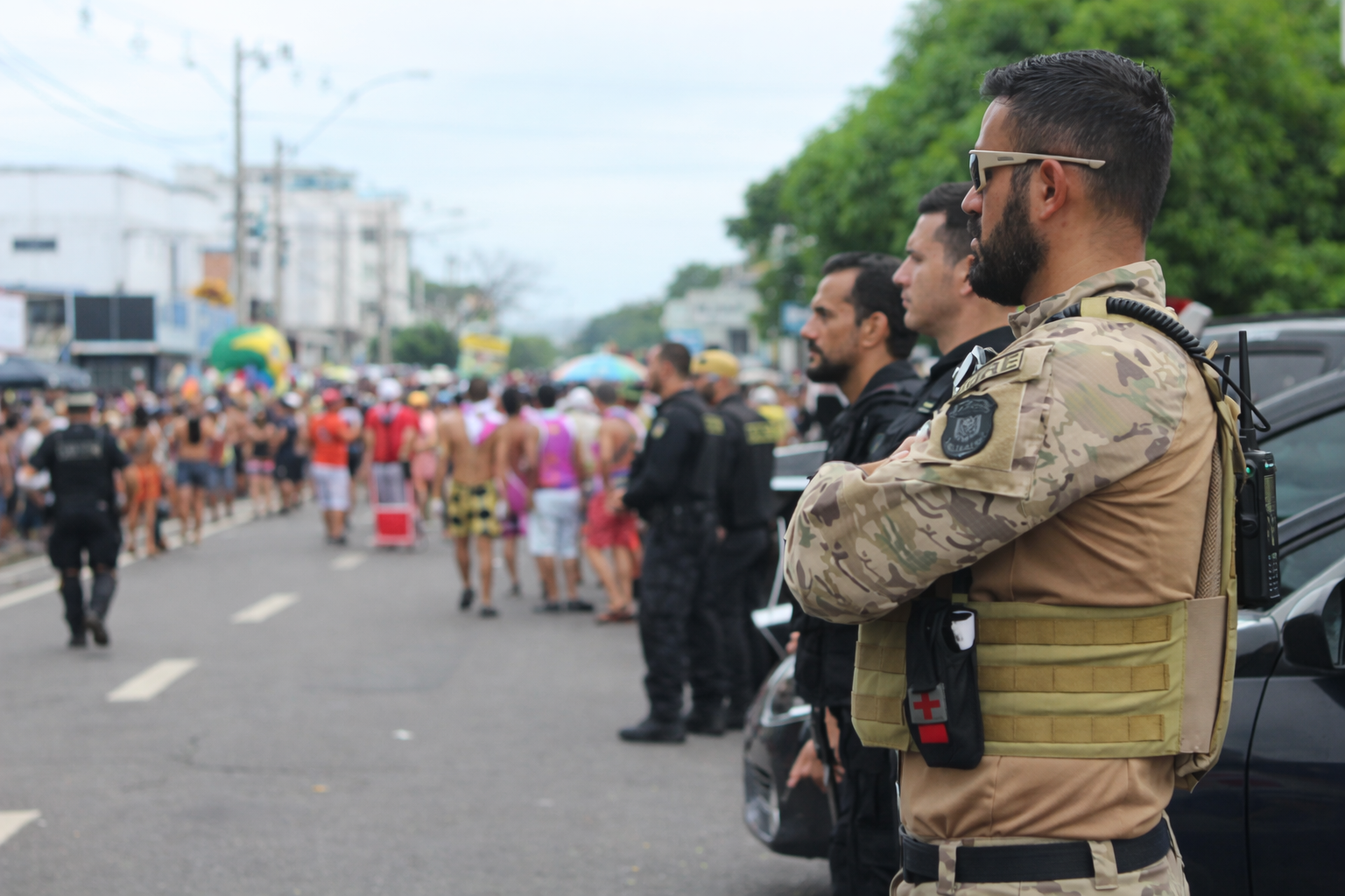 Agentes de segurança monitoram bloco de rua durante o carnaval, com viaturas ao fundo e foliões participando do evento em área urbana.