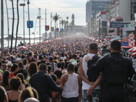 Grande público participa de bloco de carnaval na orla marítima, com prédios ao fundo e vendedores ambulantes ao longo da avenida.