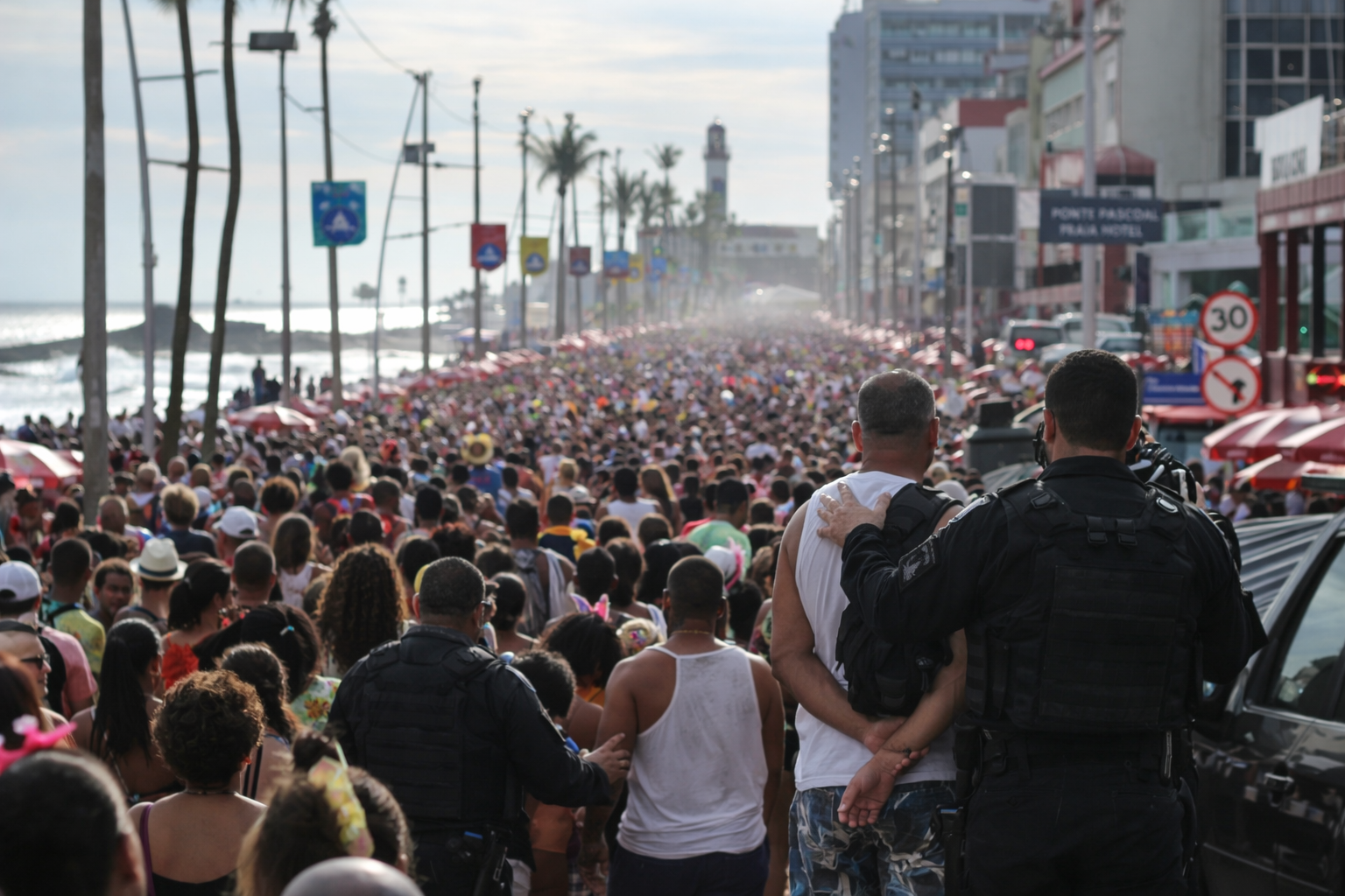 Grande público participa de bloco de carnaval na orla marítima, com prédios ao fundo e vendedores ambulantes ao longo da avenida.