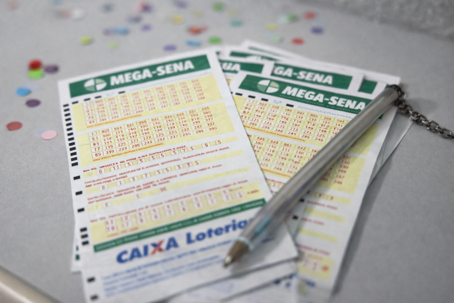 Volantes da Mega-Sena com caneta apoiada e confetes ao fundo, representando apostas em período festivo
