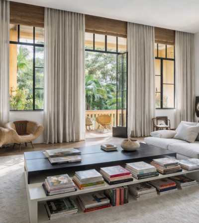 Sala de estar moderna com sofá claro, mesa de centro com livros e grandes janelas integradas à varanda com vista para área verde