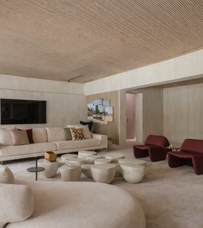 Sala de estar contemporânea com sofá curvo claro, poltronas vermelhas e mesas de centro esculturais em ambiente com teto de madeira e iluminação natural