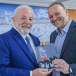 Presidente Lula e Edinho Silva sorrindo e segurando livro durante encontro em gabinete oficial com bandeira do Brasil ao fundo