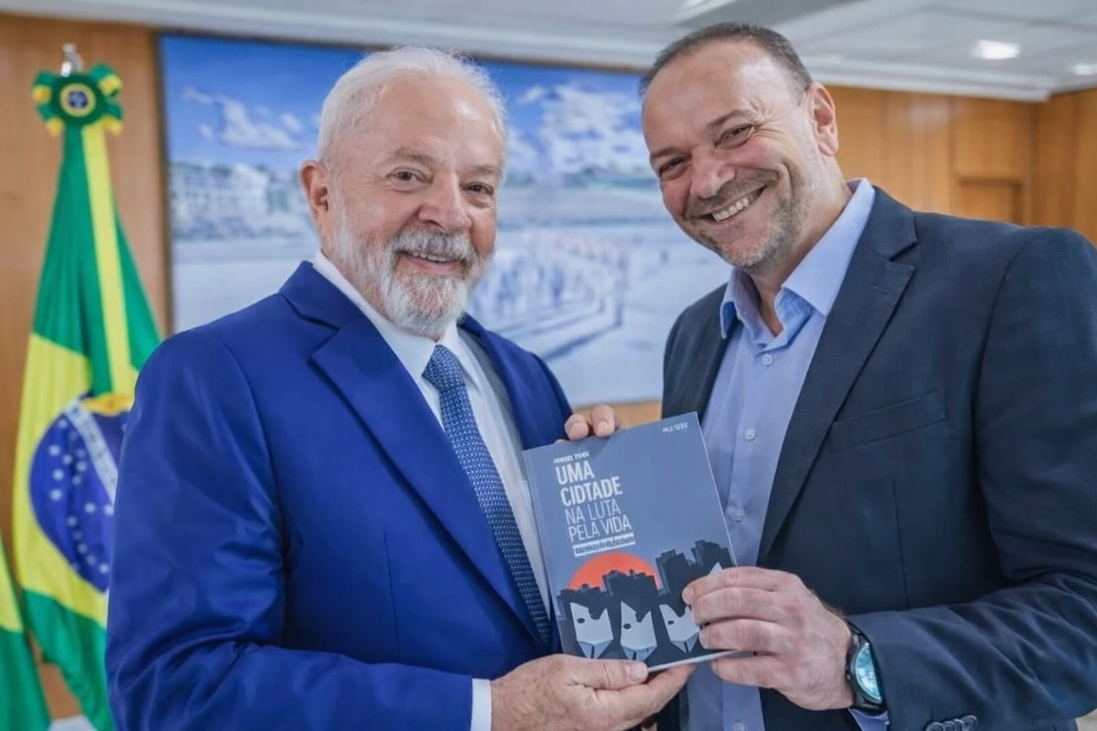 Presidente Lula e Edinho Silva sorrindo e segurando livro durante encontro em gabinete oficial com bandeira do Brasil ao fundo