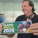 Homem sorrindo segura adesivo “Flávio Senador 2026” em ambiente interno durante período de Carnaval