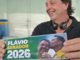 Homem sorrindo segura adesivo “Flávio Senador 2026” em ambiente interno durante período de Carnaval
