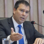 Ex-presidente do BRB fala ao microfone durante reunião oficial, usando terno escuro e gravata azul em ambiente corporativo