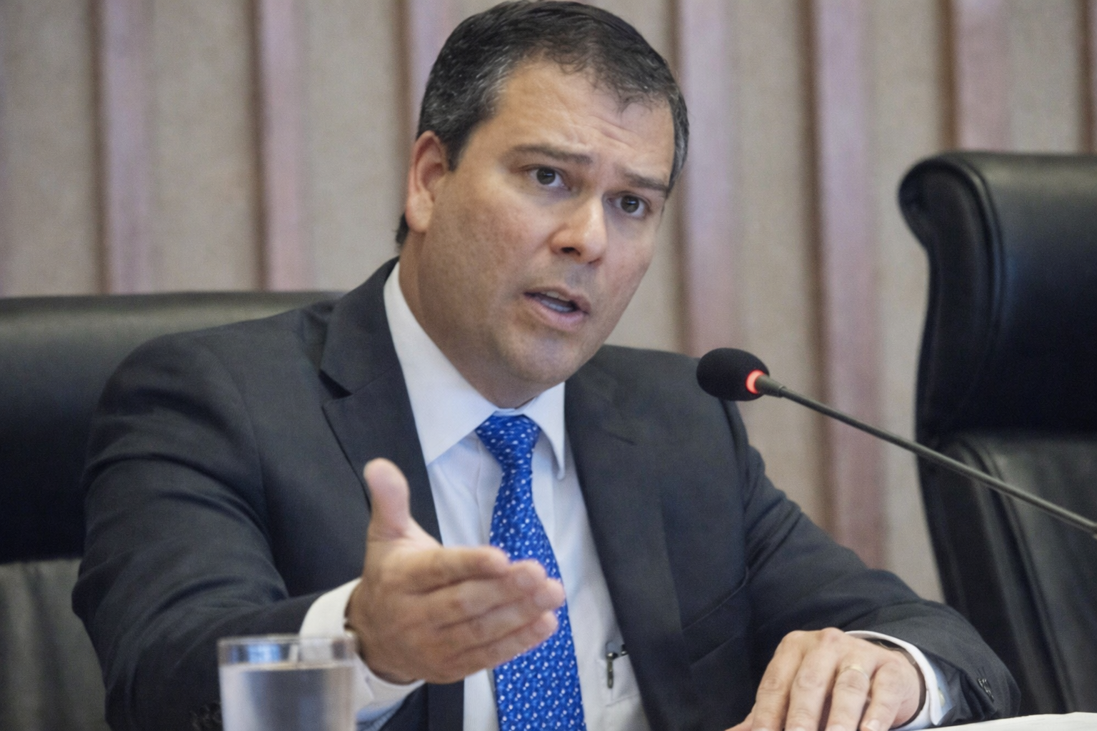 Ex-presidente do BRB fala ao microfone durante reunião oficial, usando terno escuro e gravata azul em ambiente corporativo