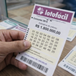 Mão segurando bilhete da Lotofácil concurso 3615 com prêmio estimado em R$ 1,8 milhão sobre balcão de lotérica.