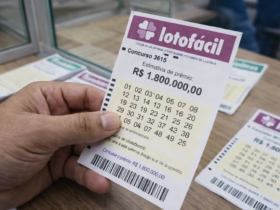 Mão segurando bilhete da Lotofácil concurso 3615 com prêmio estimado em R$ 1,8 milhão sobre balcão de lotérica.