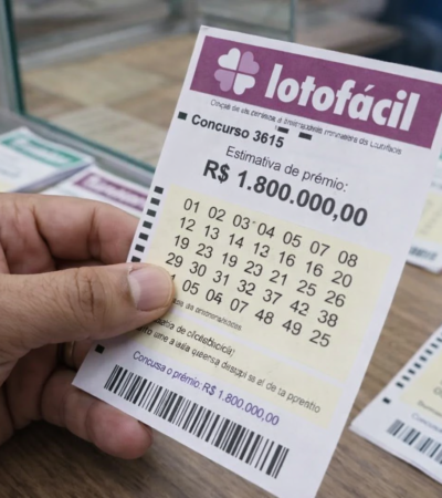 Mão segurando bilhete da Lotofácil concurso 3615 com prêmio estimado em R$ 1,8 milhão sobre balcão de lotérica.