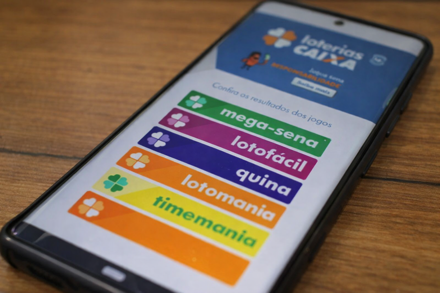 Celular com aplicativo Loterias Caixa aberto mostrando Mega-Sena, Lotofácil e Quina na tela.