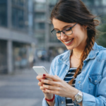 Jovem utilizando smartphone ao ar livre, sorrindo enquanto acessa aplicativo em ambiente urbano moderno.