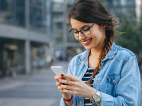 Jovem utilizando smartphone ao ar livre, sorrindo enquanto acessa aplicativo em ambiente urbano moderno.