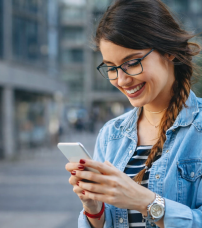 Jovem utilizando smartphone ao ar livre, sorrindo enquanto acessa aplicativo em ambiente urbano moderno.