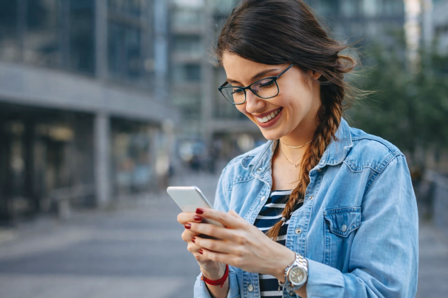 Jovem utilizando smartphone ao ar livre, sorrindo enquanto acessa aplicativo em ambiente urbano moderno.