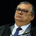 Retrato em três quartos do ministro Flávio Dino durante sessão no STF, com toga, gravata azul e fundo escuro desfocado.