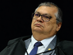 Retrato em três quartos do ministro Flávio Dino durante sessão no STF, com toga, gravata azul e fundo escuro desfocado.