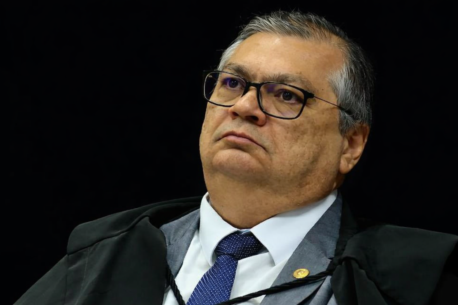Retrato em três quartos do ministro Flávio Dino durante sessão no STF, com toga, gravata azul e fundo escuro desfocado.