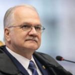 Retrato em três quartos do ministro Edson Fachin no plenário do STF, usando toga, gravata azul e sentado em cadeira marrom com microfone ao lado.